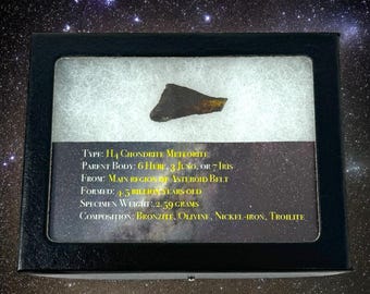 May include: A small, dark grey meteorite fragment in a black display case with a white background. The case has a label with the text "TYPE: H4 CHONDRITE METEORITE", "PARENT BODY: 6 HERE, 3 JUNO, OR 7 IRIS", "FROM: MAIN REGION OF ASTEROID BELT", "FORMED: 1.5 BILLION YEARS OLD", "SPECIMEN WEIGHT: 2.59 GRAMS", and "COMPOSITION: BRONZITE, OLIVINE, NICKEL-IRON, TROILITE".