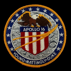 Puede incluir: Un parche bordado azul y dorado con un águila calva, el texto "APOLLO 16" y los nombres "YOUNG, MATTINGLY, DUKE" alrededor de la parte inferior.