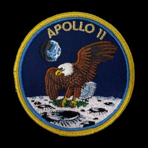 Op de afbeelding: Een ronde geborduurde patch met het embleem van de Apollo 11-missie. Het ontwerp toont een Amerikaanse zeearend die landt op het maanoppervlak, met de aarde zichtbaar in de donkerblauwe lucht. De tekst APOLLO 11 is geborduurd met gouddraad.