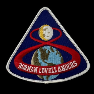 May include: A triangular patch with a blue background and white trim. The patch features a stylized Earth, a crescent moon, and a red orbital path. The text "BORMAN LOVELL ANDERS" is in white.