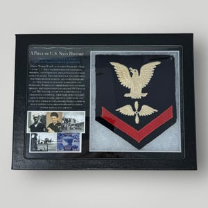 May include: A framed display featuring a vintage U.S. Navy Aviation Machinist's Mate rating badge. The badge is navy blue with a red chevron, a gold eagle, and a gold propeller. The frame includes a black background with a white inscription that reads "A Piece of U.S. Navy History."  The inscription includes a brief history of the Aviation Machinist's Mate role in the U.S. Navy during World War II.  The frame also includes three black and white photographs.