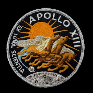 May include: Embroidered Apollo XIII mission patch. The patch features a sun, two winged horses, and the text "APOLLO XIII" and "EX LUNA, SCIENTIA" on a black background. The patch has a silver border.