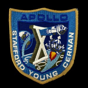 May include: Embroidered Apollo X mission patch. The patch features a blue shield shape with gold trim. The design includes a stylized "X", spacecraft, and the names STAFFORD, YOUNG, and CERNAN. The word APOLLO is at the top.