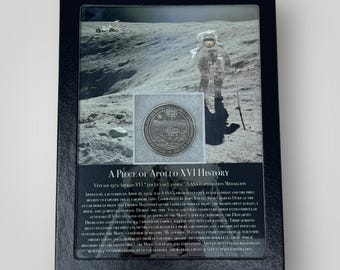 May include: A black framed display case containing a silver Apollo XVI 5th Lunar Landing NASA Exploration Medallion. The case features a black and white photograph of an astronaut on the moon. The text "A Piece of Apollo XVI History" is printed at the bottom of the case.