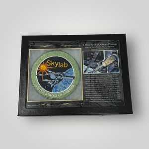 Pode incluir: Um remendo NASA Skylab preto e verde emoldurado com o texto "A Piece of NASA Space History" e "Original Space Station Skylab Program Astronaut Mission Patch".