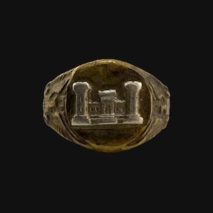 Puede incluir: Un anillo vintage dorado con un diseño en relieve de un castillo. El anillo tiene una cara ovalada con la imagen del castillo en el centro. La banda del anillo tiene un diseño texturizado y detallado.