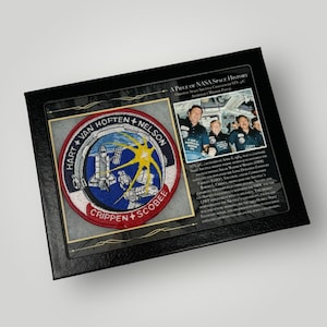 May include: A framed black and gold display with a NASA space shuttle mission patch. The patch is red, white, and blue with the names of the astronauts: Hart, Van Hoften, Nelson, Crippen, and Scobee. The text "A Piece of NASA Space History" is printed on the frame.