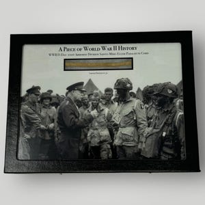 Könnte beinhalten: Ein gerahmtes Schwarzweißfoto von Soldaten während des Zweiten Weltkriegs. Das Foto wird von einem Stück Fallschirmleine aus der Invasion am D-Day begleitet. Der Text "A Piece of World War II History" und "WWII D-Day 101st Airborne Division Sainte-Mère-Église Parachute Cord" ist auf dem Rahmen gedruckt.