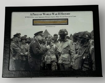 May include: A framed black and white photograph of soldiers during World War II. The photo is accompanied by a piece of parachute cord from the D-Day invasion. The text "A Piece of World War II History" and "WWII D-Day 101st Airborne Division Sainte-Mère-Église Parachute Cord" is printed on the frame.
