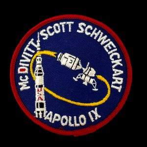 May include: A circular patch with a blue background and a red border. The patch features a rocket, a spacecraft, and the text "McDivitt/Scott Schweickart" and "Apollo IX". The word "USA" is also visible. The design is embroidered with white and yellow thread.