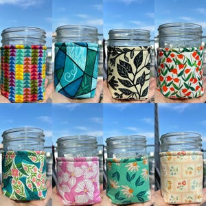 May include: A collection of eight glass mason jars, each adorned with a unique fabric sleeve. The sleeves feature various colorful patterns, including geometric shapes, floral designs, and botanical prints. The jars are displayed against a bright blue sky.