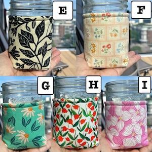 K&ouml;nnte beinhalten: F&uuml;nf Einmachgl&auml;ser aus Glas, jedes mit einer anderen Stoffh&uuml;lle umwickelt. H&uuml;lle E zeigt schwarze Bl&auml;tter auf cremefarbenem Grund. F hat ein Gitter aus Blumenbriefmarken. G zeigt wei&szlig;e G&auml;nsebl&uuml;mchen auf T&uuml;rkis. H zeigt rote Blumen auf Wei&szlig;, I zeigt rosa Blumen.