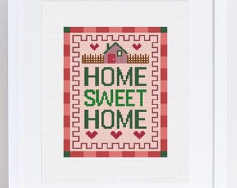 Op de afbeelding: Een ingelijste kruissteekafbeelding. Het ontwerp toont een huis, harten en de woorden "HOME SWEET HOME" in groen. De achtergrond heeft een rood-roze geruit patroon.