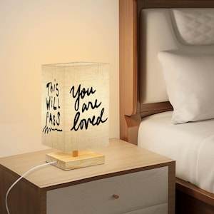 May include: A rectangular bedside lamp with a linen shade and a wooden base. The shade displays the text "This Will Pass" and "You are loved" in black script. The lamp is plugged into a wooden nightstand.