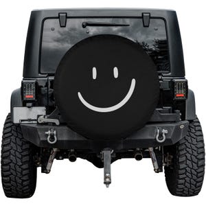 K&ouml;nnte beinhalten: Schwarze Reserveradabdeckung mit einem wei&szlig;en Smiley-Gesicht. Die Abdeckung befindet sich auf einem schwarzen Jeep.