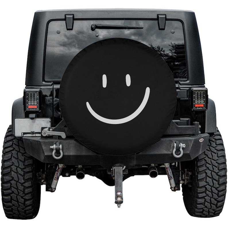 K&ouml;nnte beinhalten: Schwarze Reserveradabdeckung mit einem wei&szlig;en Smiley-Gesicht. Die Abdeckung befindet sich auf einem schwarzen Jeep.