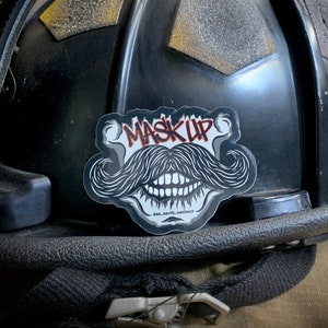 Peut inclure: Un casque de pompier noir avec un autocollant blanc qui dit "MASK UP" en lettres rouges. L'autocollant représente un crâne stylisé avec une moustache et des dents.