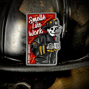 Peut inclure: Un autocollant représentant un pompier squelette en uniforme noir, tenant une tasse de café et fumant un cigare. L'autocollant porte l'inscription "Smells Like Work" sur un fond de briques rouges. Il y a aussi le texte "EN ROUTE GRAPHICS".