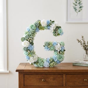 May include: A large floral letter "G" made of blue, green, and white flowers, including roses and hydrangeas. The letter sits on a wooden table with a drawer. A framed botanical print is in the background.
