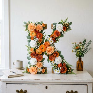 May include: A wooden letter 'K' adorned with orange, white, and yellow flowers, including roses and daisies. The floral display is set against a white wall, with a small vase of flowers and a coffee cup on a white dresser.