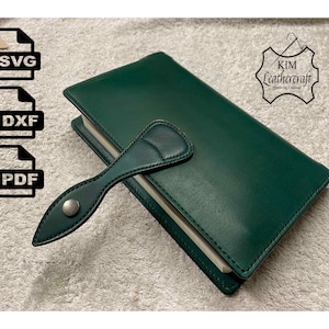 May include: A dark green leather-bound journal with a matching leather strap and a silver metal button. The journal is closed and has a white spine. The journal is on a white fluffy surface. The text "KIM Leathercraft" is visible in the top right corner of the image.