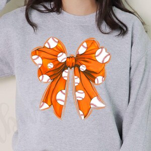 May include: A gray sweatshirt with an orange bow featuring a white baseball pattern.