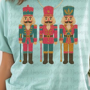 May include: A light blue t-shirt featuring three colorful nutcracker soldier designs. Each nutcracker has a different uniform color, including red, teal, and gold, with black boots and a matching hat. The shirt has the text "Delighted Business" repeated.