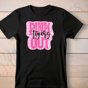 May include: Black t-shirt with a pink and white design that reads "PINK tigers OUT" in a playful font. The text is inside a pink, irregular-shaped box. The shirt is made of a soft material.