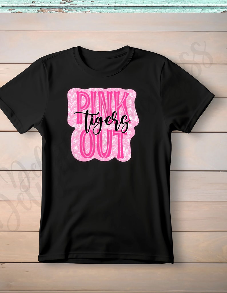 May include: Black t-shirt with a pink and white design that reads "PINK tigers OUT" in a playful font. The text is inside a pink, irregular-shaped box. The shirt is made of a soft material.