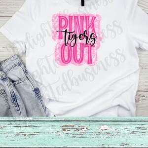 May include: White t-shirt with a pink and black graphic. The graphic reads "PINK tigers OUT" in a combination of bold and script fonts. The shirt is paired with light wash denim jeans.