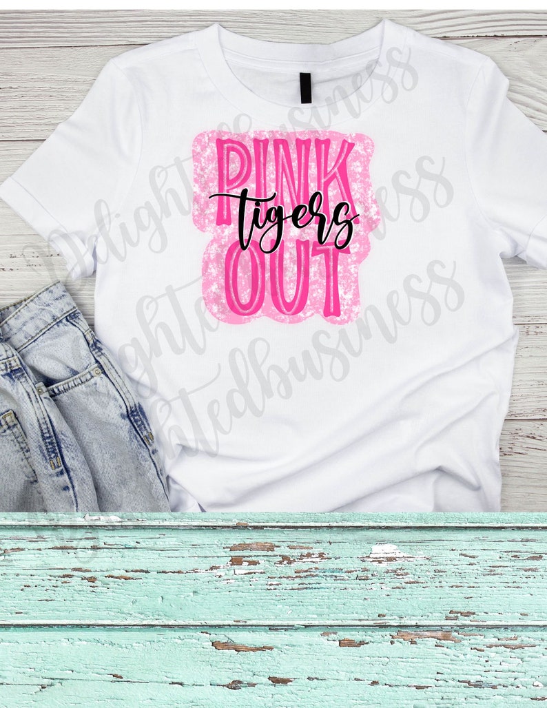 May include: White t-shirt with a pink and black graphic. The graphic reads "PINK tigers OUT" in a combination of bold and script fonts. The shirt is paired with light wash denim jeans.