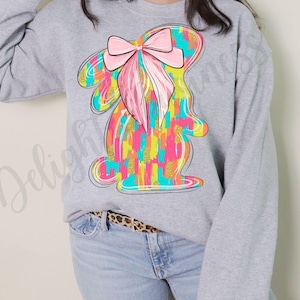 May include: A gray sweatshirt with a colorful abstract bunny design. The bunny has a large pink bow on its head. The design is made up of bright colors, including yellow, pink, blue, and green.