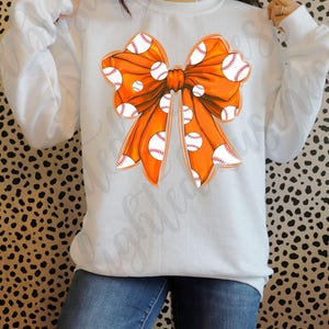May include: A white sweatshirt with an orange bow design. The bow is decorated with white baseballs.