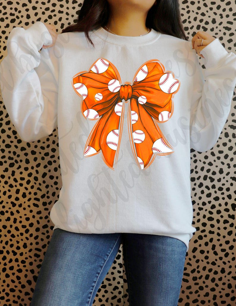 May include: A white sweatshirt with an orange bow design. The bow is decorated with white baseballs.