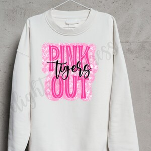 May include: Cream-colored sweatshirt with the words "PINK TIGERS OUT" in a pink, textured design. The word "tigers" is written in a black script font. The sweatshirt is hanging on a white hanger.