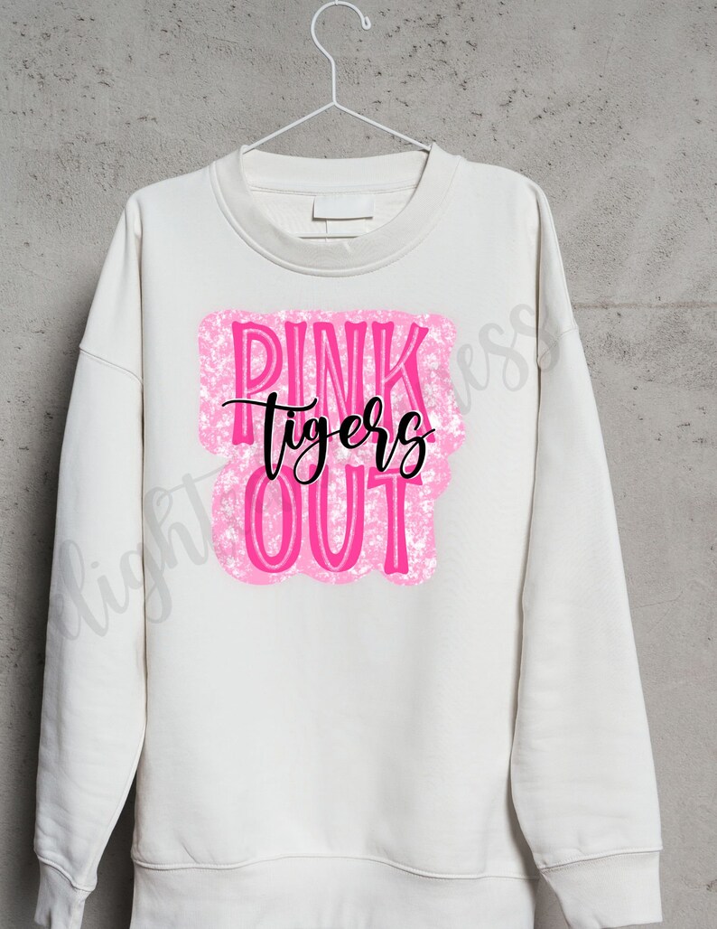May include: Cream-colored sweatshirt with the words "PINK TIGERS OUT" in a pink, textured design. The word "tigers" is written in a black script font. The sweatshirt is hanging on a white hanger.