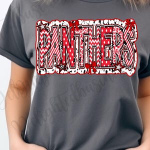 May include: A gray t-shirt featuring the word "PANTHERS" in a bold, decorative design. The text is primarily red with white accents, including polka dots, stripes, and zigzags. The design is outlined in black and white, with star and bow details.