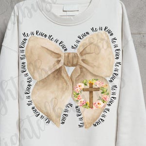 Puede incluir: Una sudadera blanca con un lazo marrón y una corona de flores con una cruz en el centro. El texto "He is Risen" se repite alrededor del lazo.
