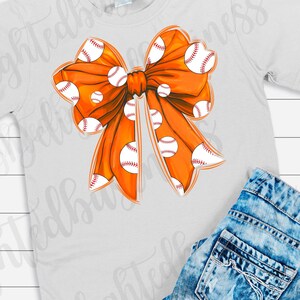May include: Orange bow with white baseballs printed on it. The bow is on a white t-shirt.
