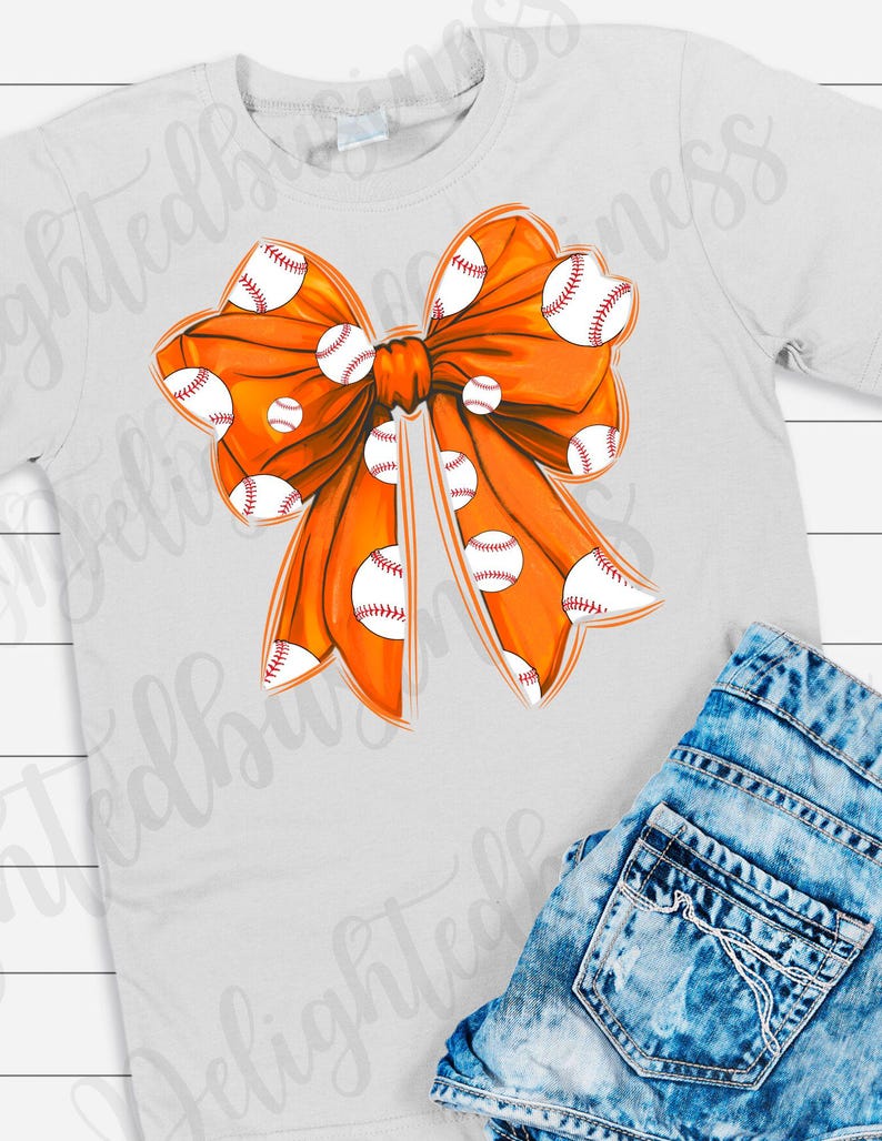 May include: Orange bow with white baseballs printed on it. The bow is on a white t-shirt.