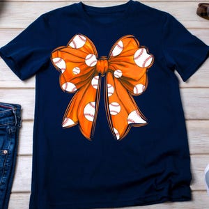 May include: A navy blue t-shirt with an orange bow decorated with white baseballs.