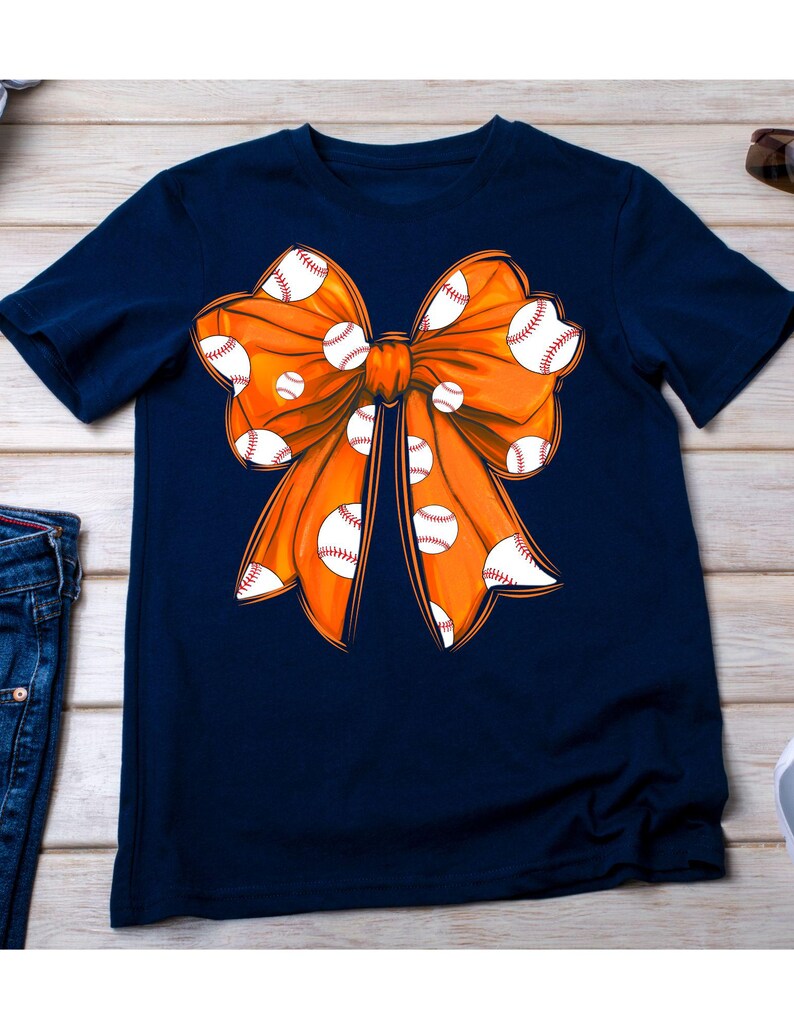 May include: A navy blue t-shirt with an orange bow decorated with white baseballs.