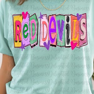 Puede incluir: Una camiseta azul claro con el texto "RED DEVILS" en un diseño de letras de bloque 3D colorido. Las letras están delineadas en negro y decoradas con corazones y estrellas.