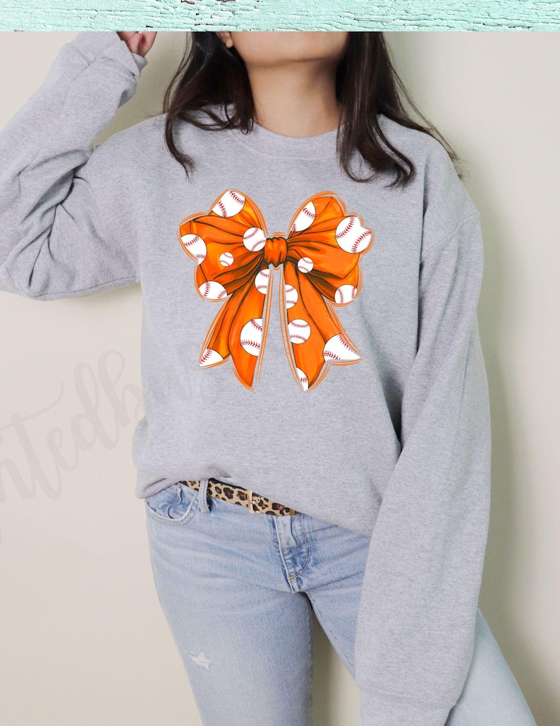 May include: A gray sweatshirt with an orange bow featuring a white baseball pattern.