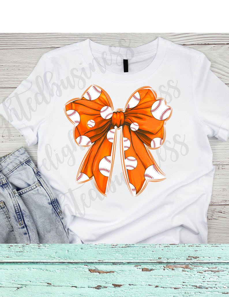 May include: Orange bow with white baseballs printed on it. The bow is on a white t-shirt.