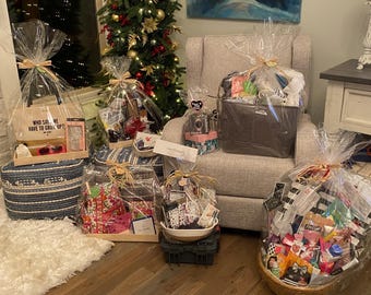 May include: Several gift baskets wrapped in clear cellophane are arranged on a wooden floor. The baskets contain various items, including a tote bag with text, sunglasses, and other gifts. A Christmas tree with gold ornaments is in the background.