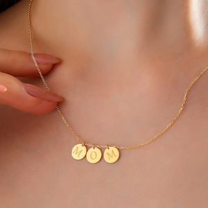 May include: A gold-toned necklace featuring a delicate chain and three small, circular pendants. Each pendant is engraved with a single letter, collectively spelling the word MOM in a clean, sans-serif font centered along the collarbone area.