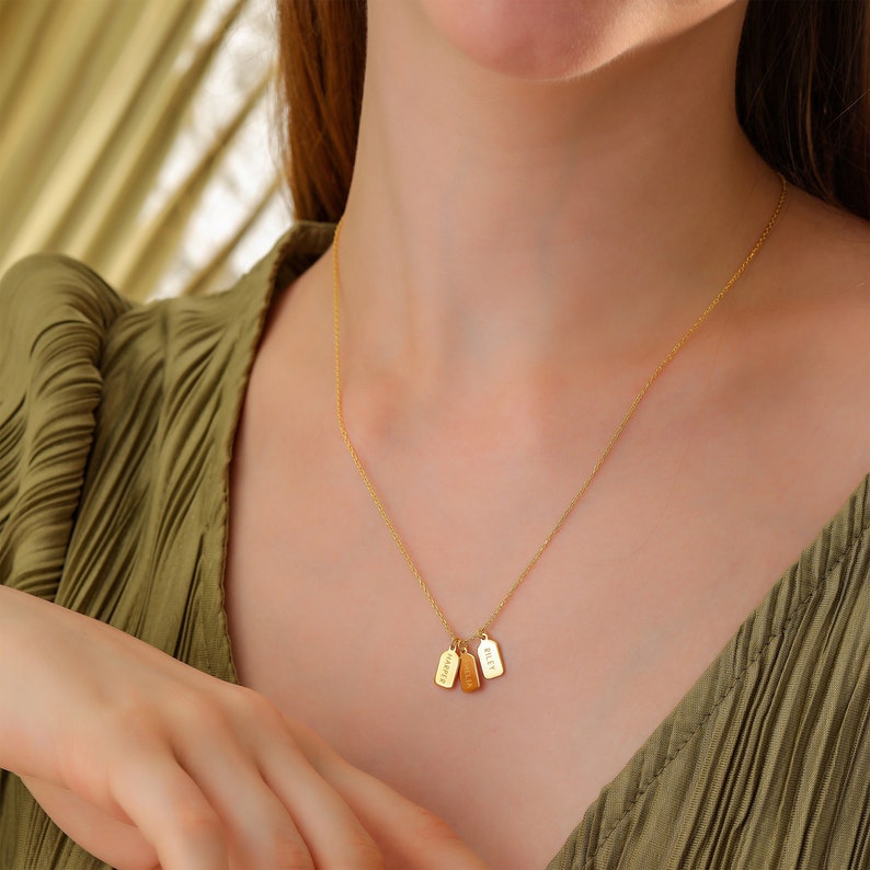 A woman wearing a necklace with three mini tags poses with her head tilted to the side