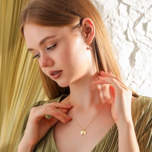 A woman wearing a 14k solid gold necklace with three mini tags looks down as she adjusts the necklace