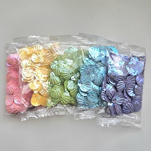 Peut inclure: Cinq sachets en plastique transparent contenant des sequins en forme de coquillage dans un dégradé de couleurs incluant rose, jaune, vert clair, bleu clair et violet. Chaque sequin présente une texture nervurée et deux petits trous.
